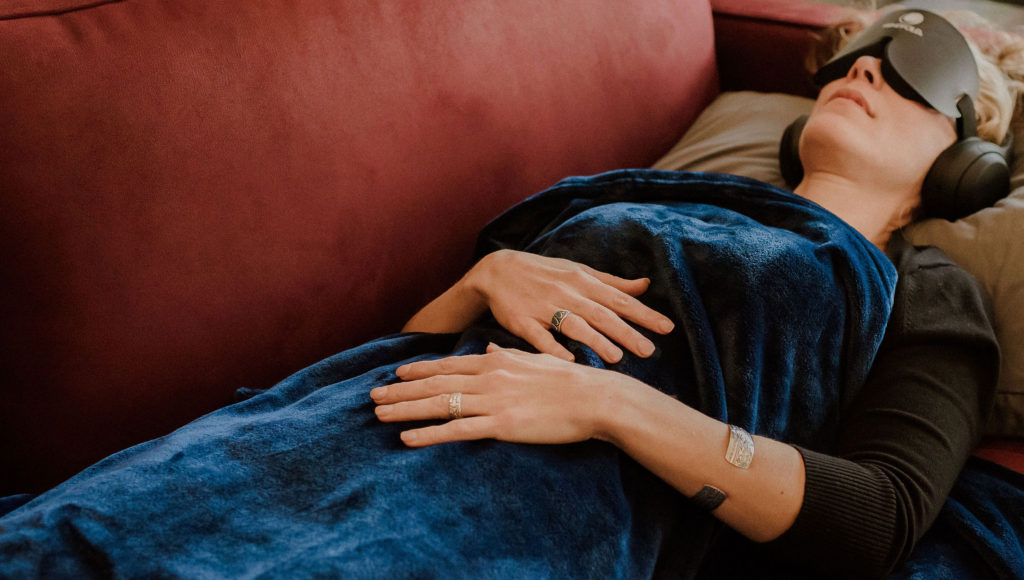 woman laying on couch wearing headphones and covered with a blanket participating in psychedelic therapy